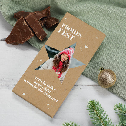 Weihnachtsschokolade mit Foto im Stern und individuellem Text. Verpackung in Kraftpapieroptik mit weißen Sternen. Im Ausschnitt ein winterliches Porträt einer Frau mit Mütze und Schal. Dekoriert mit grüner Stoffunterlage, Schokoladenstücken, goldener Kugel und Tannenzweig.
