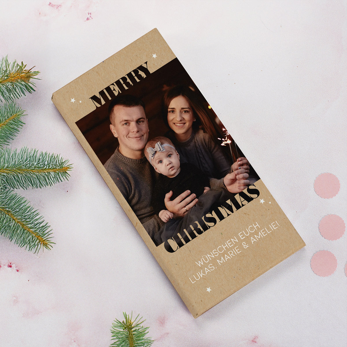 Foto-Schokoladentafel zu Weihnachten mit persönlichem Druck von Foto und Text. Auf der Verpackung ist eine junge Familie mit Kleinkind abgebildet, eingerahmt vom Schriftzug „Merry Christmas“ und kleinen weißen Sternen. Die Schokolade liegt auf einem weihnachtlich dekorierten Hintergrund mit Tannenzweigen und schafft eine warme, festliche Atmosphäre.