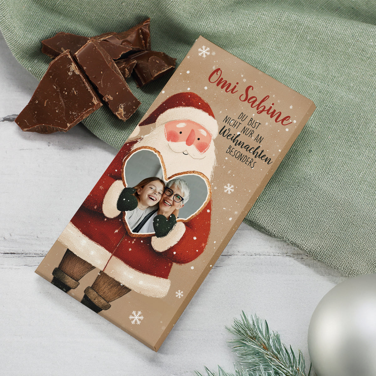 Foto-Schokoladentafel mit personalisierter Verpackung im Weihnachtsdesign. Der Weihnachtsmann hält ein Herz mit integriertem Foto, darüber steht „Omi Sabine“. Das Motiv ist von Schneeflocken umgeben, auf dem Tisch liegen Schokoladenstücke, Tannenzweige und eine silberne Kugel. Ideal als Geschenk zu Weihnachten oder Nikolaus.