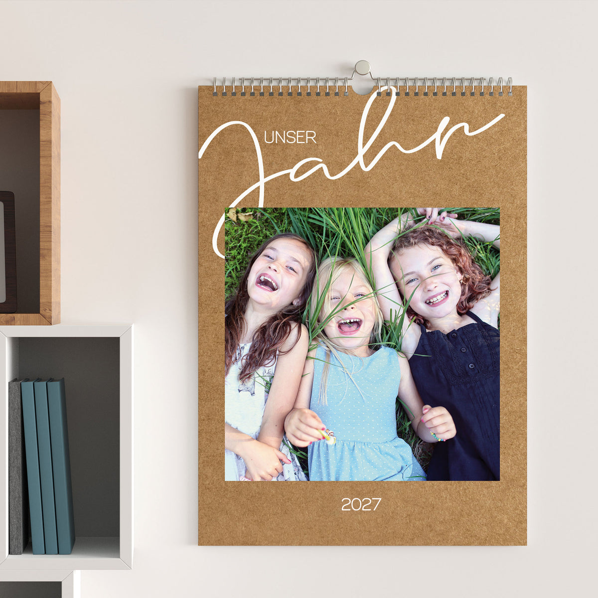 Foto-Wandkalender 2027 in Kraftpapier-Optik, aufgehängt an einer hellen Wand, mit Foto von drei lachenden Kindern und dem Titel „Unser Jahr“.