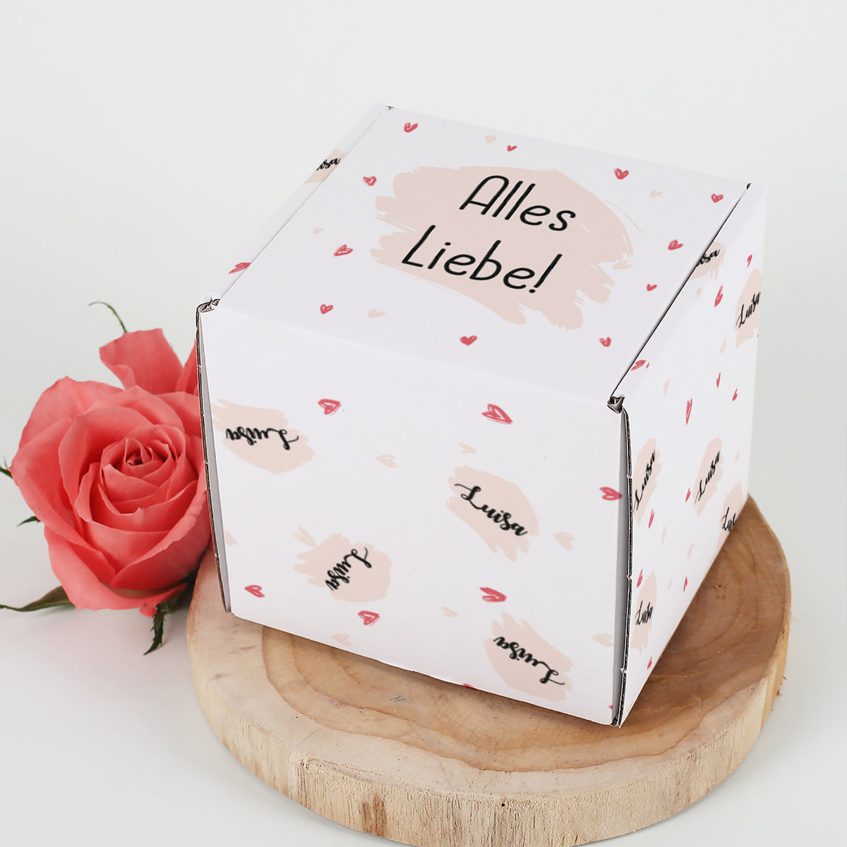 Geschenkbox mit Herz-Motiv personalisiert mit Name & Wunschtext auf einer Holzscheibe neben einer Rose fotografiert.