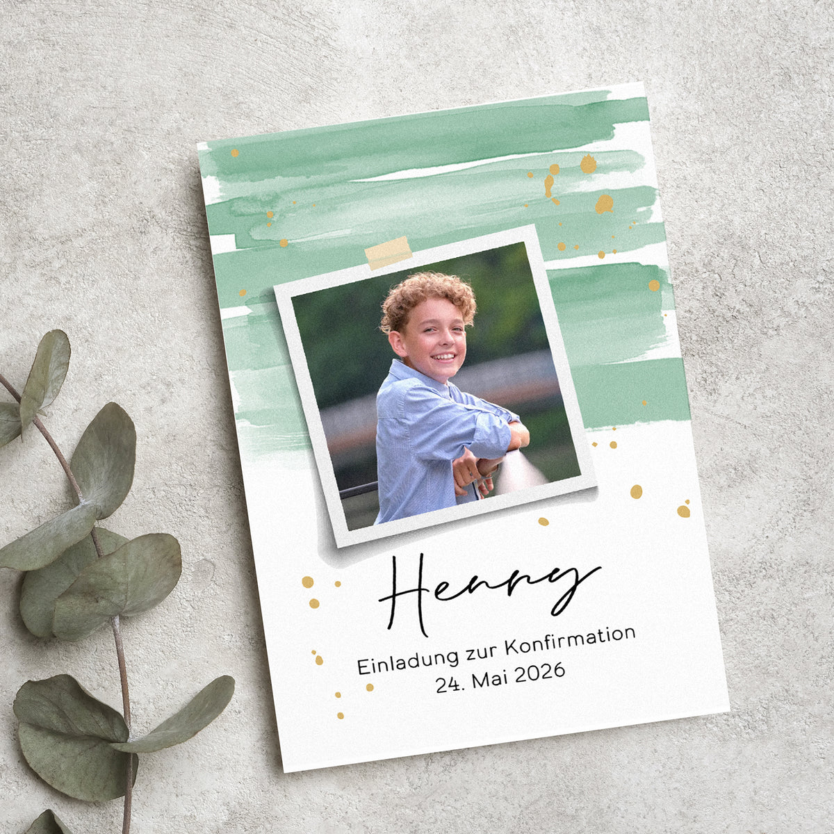 Personalisierte Einladungskarte zur Konfirmation mit modernem Aquarell-Design in Grün und Gold. Ein Polaroid-Foto des Konfirmanden ist mittig platziert, darunter sein Name und das Datum der Feier. Die Karte liegt auf einer strukturierten Steinoberfläche, umgeben von Eukalyptusblättern.