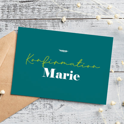 Personalisierte Einladungskarte zur Konfirmation in stilvollem Petrolgrün. Die Vorderseite trägt den Schriftzug „Konfirmation Marie“ in eleganter Schrift mit einer kleinen Ziergrafik darüber. Die Karte liegt auf einem weißen Holztisch mit einem braunen Briefumschlag und filigranen Trockenblumen, passend zur natürlichen Gestaltung.
