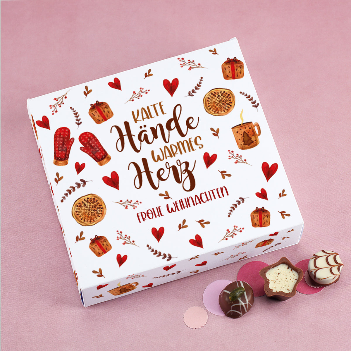 Personalisierbare Lindt Pralinenbox mit Aufdruck „Kalte Hände, warmes Herz – Frohe Weihnachten“, weihnachtlich dekoriert mit Herzen, Geschenken und Gebäck auf rosa Hintergrund.