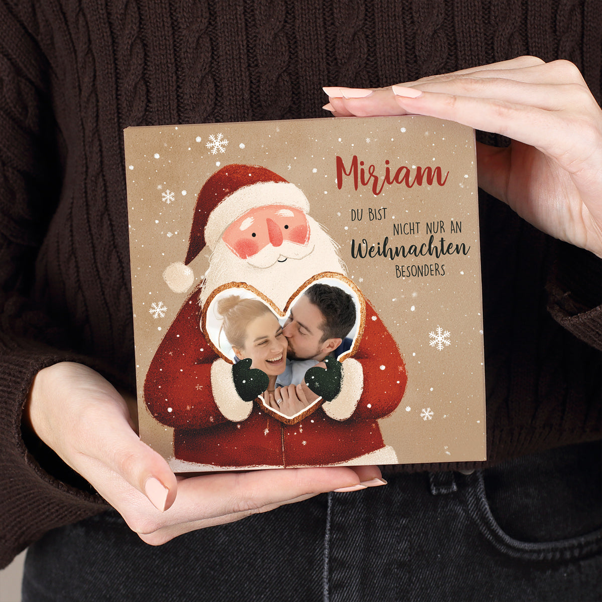 Weihnachtsmann mit freundlichem Gesichtsausdruck, hält Herz mit Foto eines Paares. Darüber personalisierte Beschriftung in roter und schwarzer Schrift. Deutlich sichtbare Struktur des Kartonmaterials und winterliches Schneeflocken-Design.