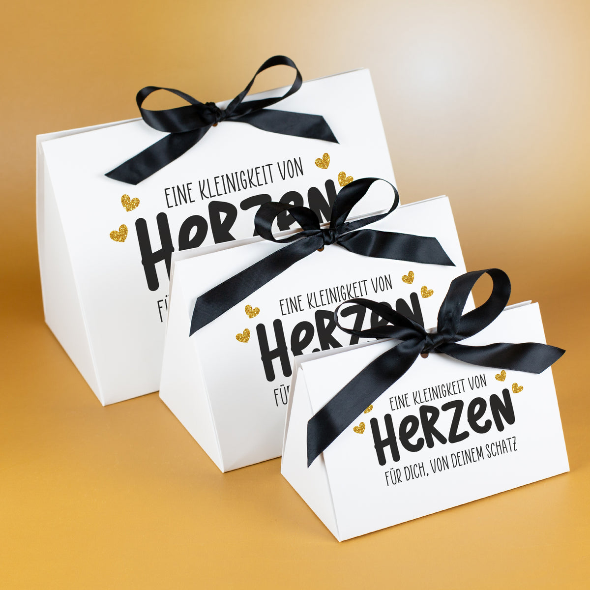 Drei Geschenkverpackungen „Eine Kleinigkeit von Herzen“ in drei Größen mit schwarzer Satinschleife auf goldenem Hintergrund
