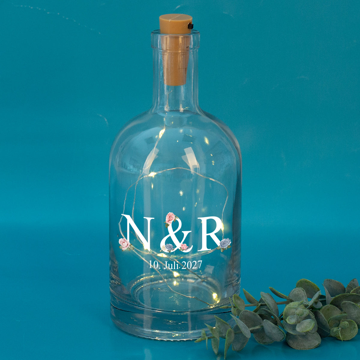 LED-Flasche mit Initialen N & R und Datum 10. Juli 2027, vor türkisfarbenem Hintergrund mit Eukalyptus