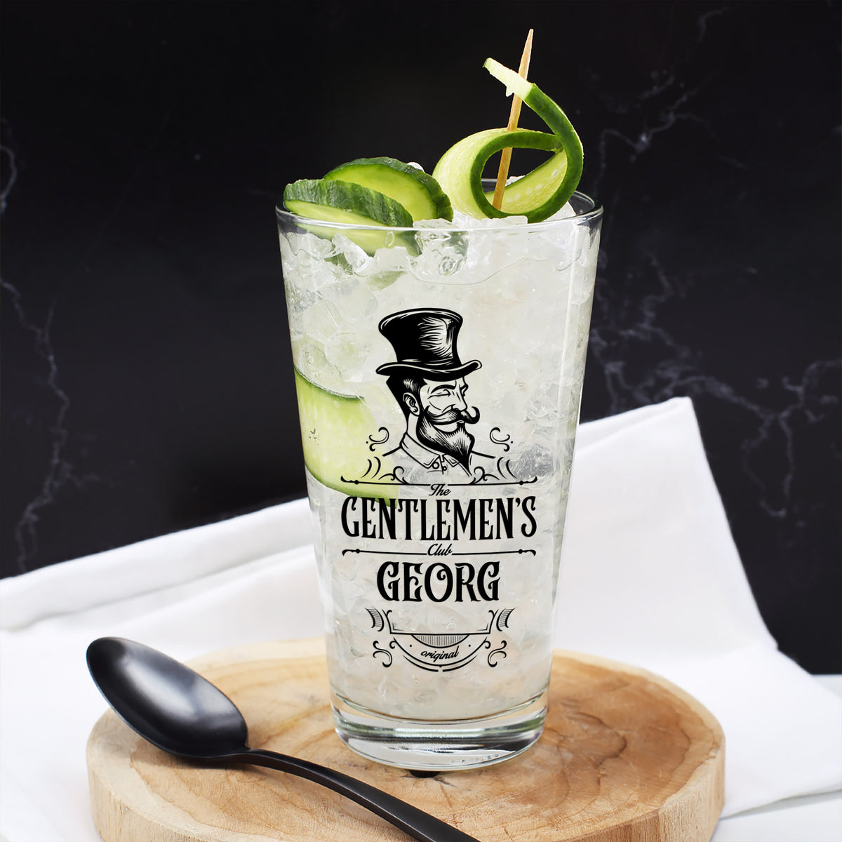 Trinkglas The Gentlemen’s Club mit Name Georg, befüllt mit Eis, Gurke und Drink, auf Holzteller mit schwarzem Löffel