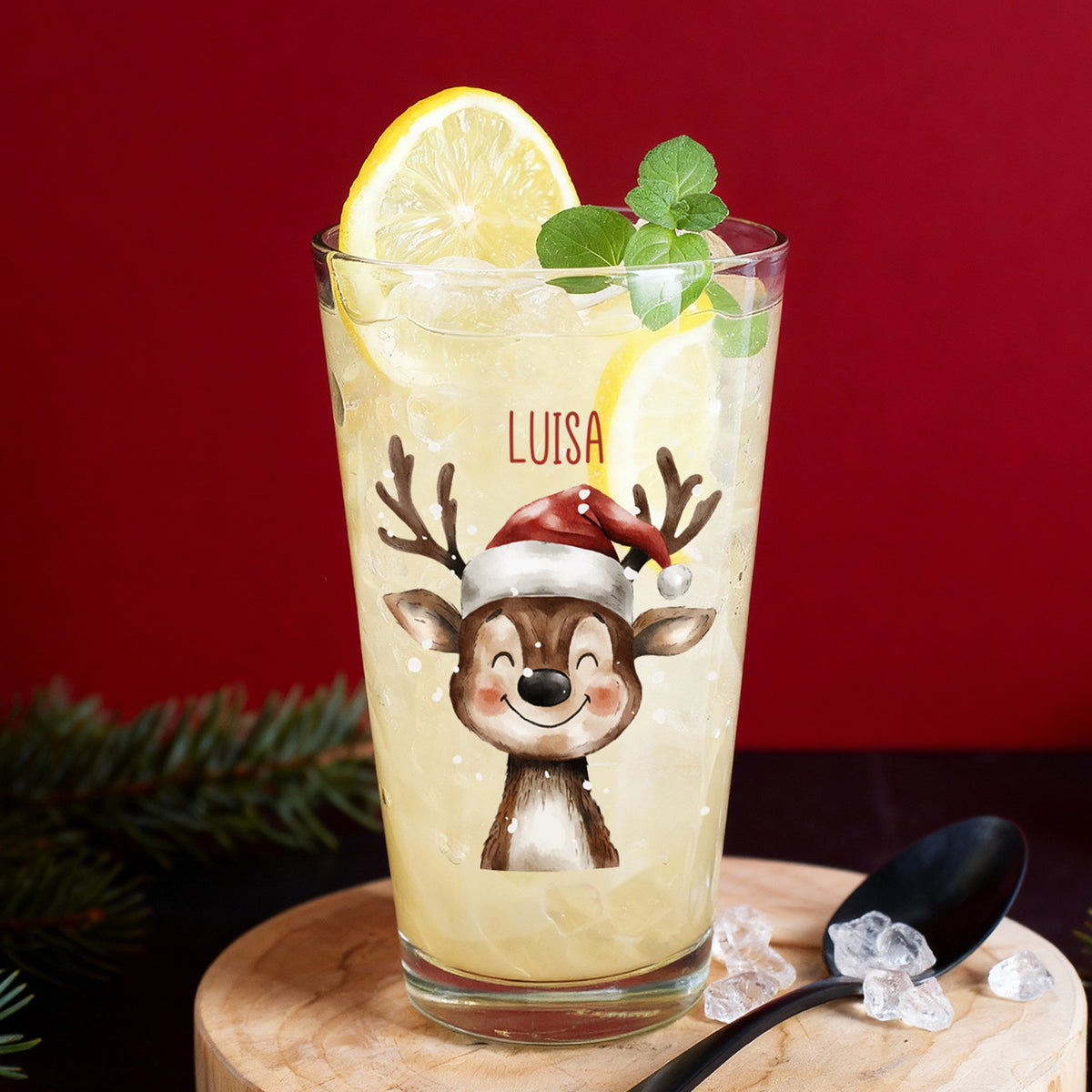 Personalisierbares Weihnachts-Trinkglas mit Rentiermotiv und Name Luisa, befüllt mit Zitronenlimonade, auf rotem Hintergrund dekoriert mit Tannenzweigen.