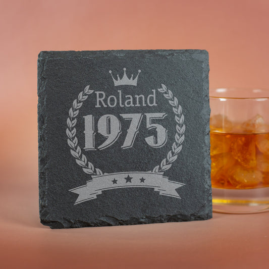 Personalisierter Schiefer-Untersetzer mit Name und Geburtsjahr - aufgestellt mit Glas im Hintergrund