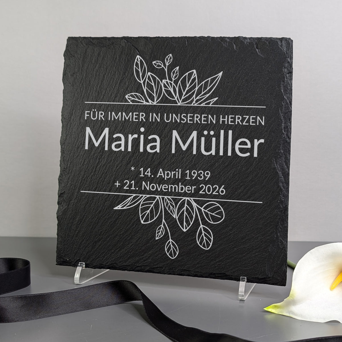 Gedenktafel auf einem Ständer dekoriert – neben Blume und Trauerband platziert