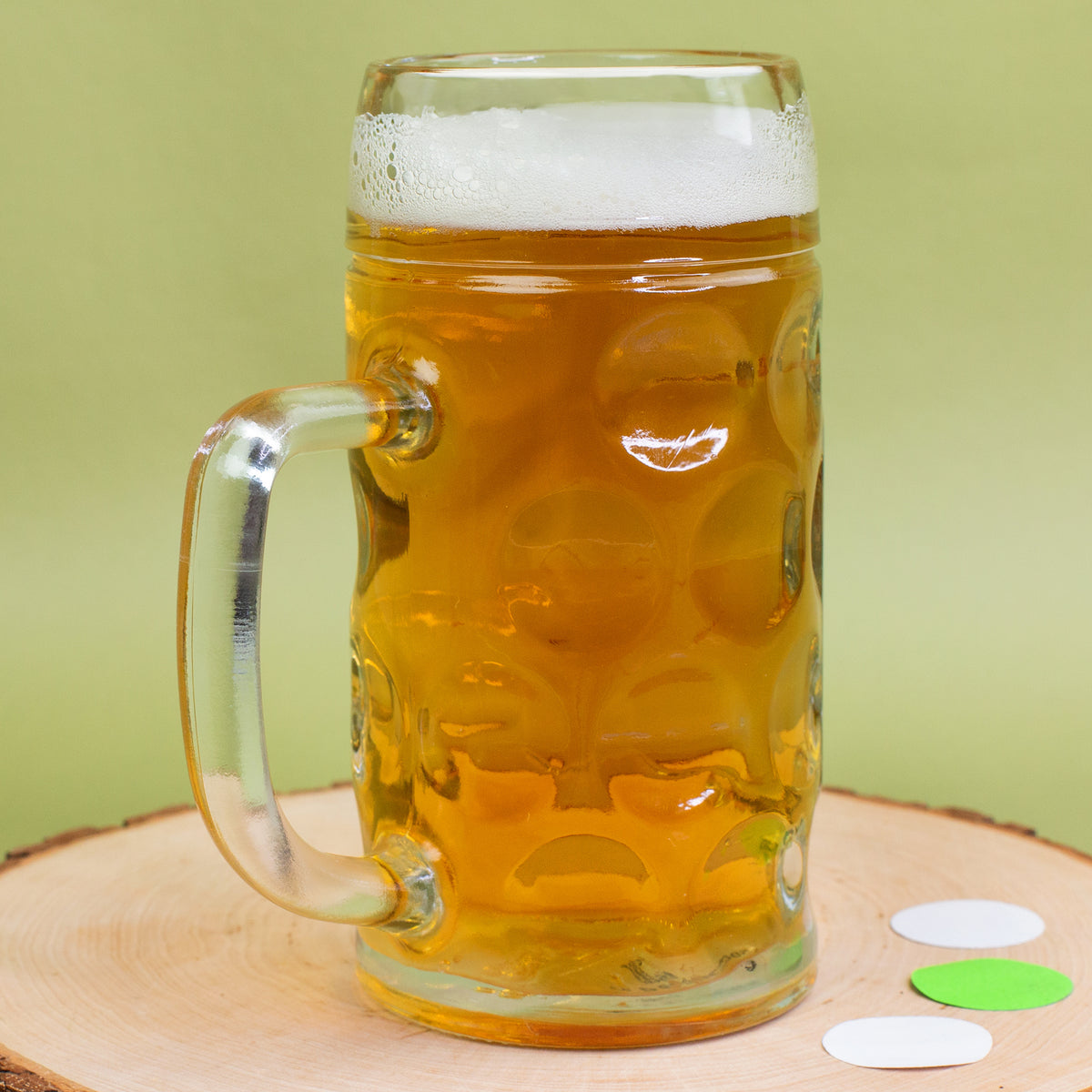 Unbedrucktes Bierglas mit Henkel, Schaumkrone und Konfetti-Deko auf Holzplatte vor grünem Hintergrund