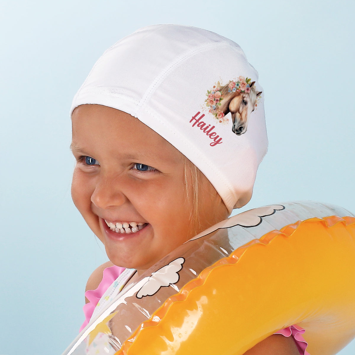 Mädchen mit Badekappe mit Pferdemotiv und Name „Hailey“ – lächelnd mit Schwimmring
