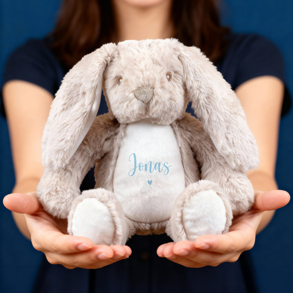 Eine Person hält einen beigefarbenen Plüschhasen mit langen Schlappohren in beiden Händen. Auf dem weißen Bauch ist der Name „Jonas“ in blauer Schrift über einem kleinen Herzmotiv aufgedruckt. Der Hintergrund ist in einem warmen Blauton gehalten.