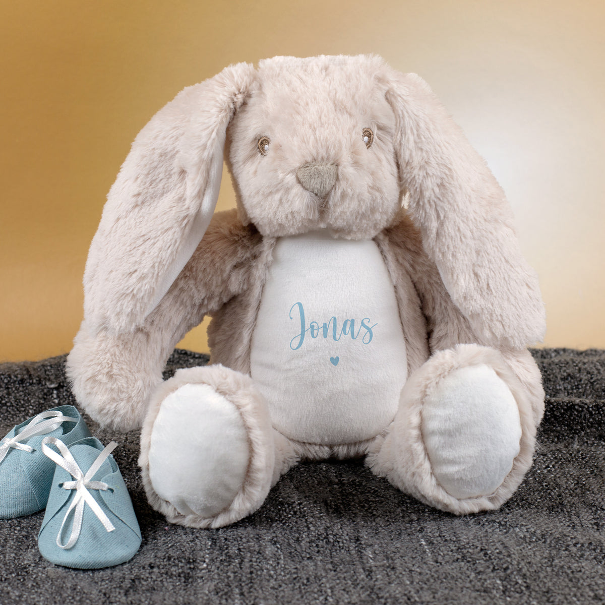 Kuscheltier-Hase in Beige mit blauer Personalisierung „Jonas“ und Herzmotiv sitzt auf einer grauen Decke. Daneben liegen kleine blaue Babyschuhe, der Hintergrund ist goldfarben.