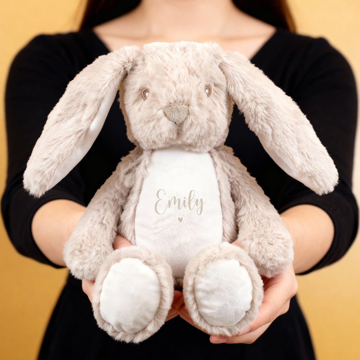 Eine Person hält einen flauschigen beigefarbenen Hasen mit der Aufschrift „Emily“ und kleinem beigem Herzmotiv auf weißem Bauch. Das Kuscheltier hat lange Ohren und einen liebevollen Gesichtsausdruck.