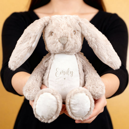 Eine Person hält einen flauschigen beigefarbenen Hasen mit der Aufschrift „Emily“ und kleinem beigem Herzmotiv auf weißem Bauch. Das Kuscheltier hat lange Ohren und einen liebevollen Gesichtsausdruck.