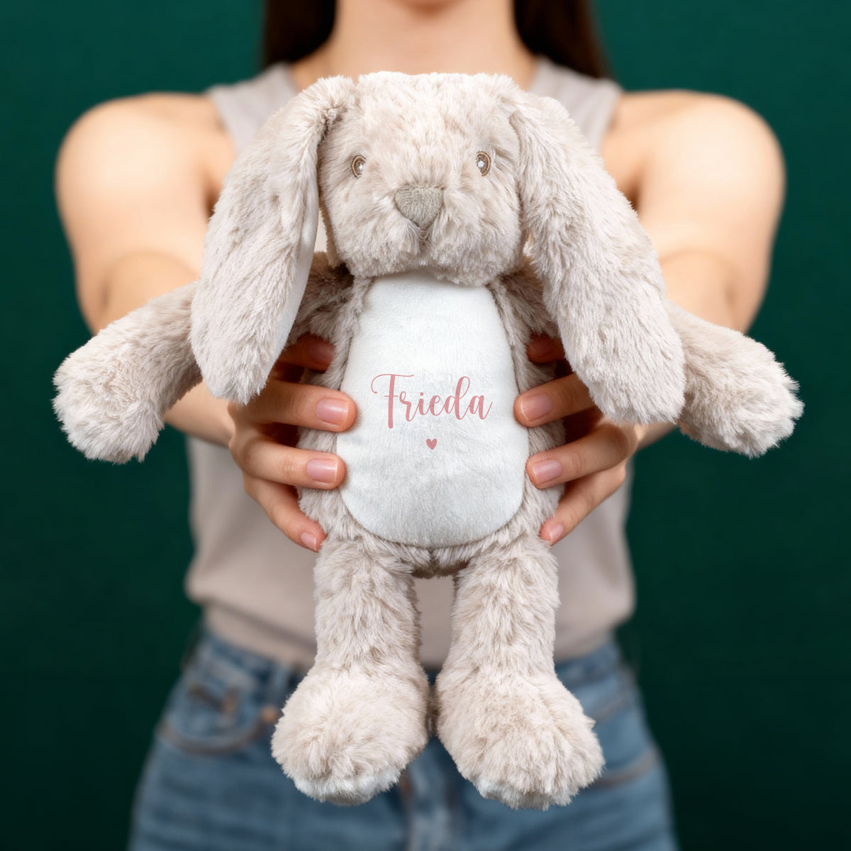 Eine Person hält einen beigefarbenen Hasen mit rosa Schriftzug „Frieda“ und Herzmotiv auf weißem Bauch vor einem dunkelgrünen Hintergrund. Der Hase wirkt weich und liebevoll gestaltet.