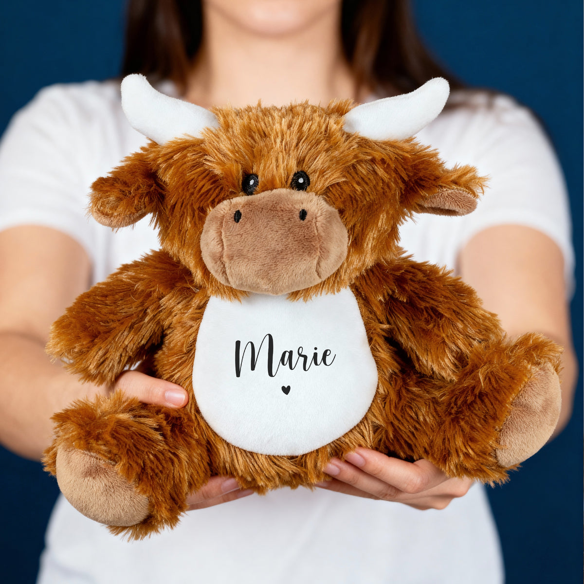 Frau hält flauschige Kuschelkuh mit Name Marie und Herzmotiv in den Händen vor dunklem Hintergrund.
