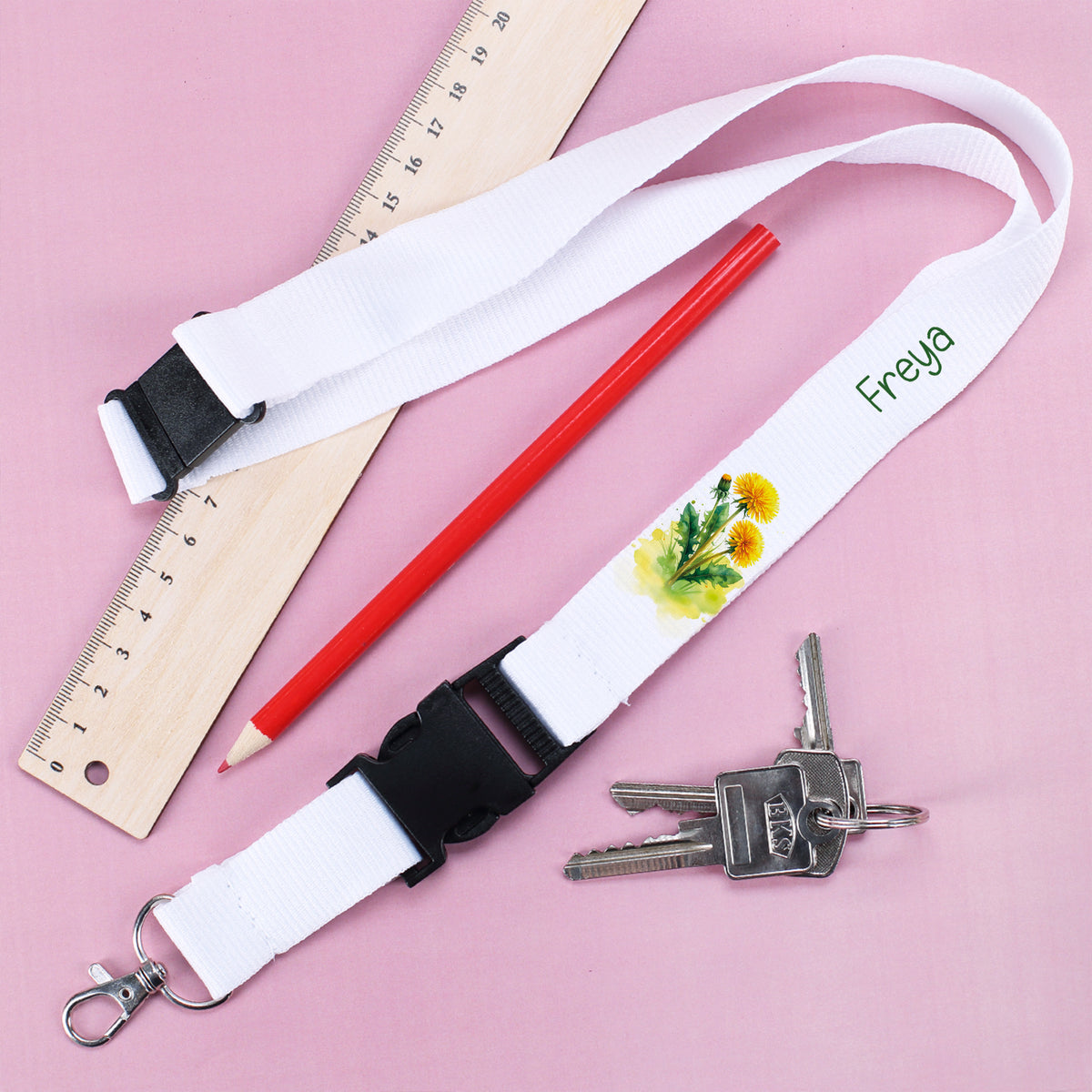 Schlüsselband mit Aufdruck "Freya" und Löwenzahn-Motiv, daneben Schlüssel, Buntstift und Lineal auf rosa Untergrund