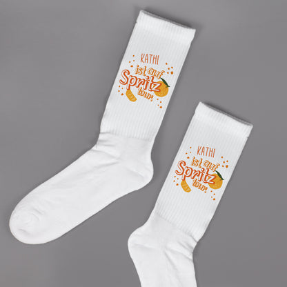 Flach liegende weiße Socken mit Spritz-Tour-Motiv und personalisiertem Namen, Studioaufnahme.