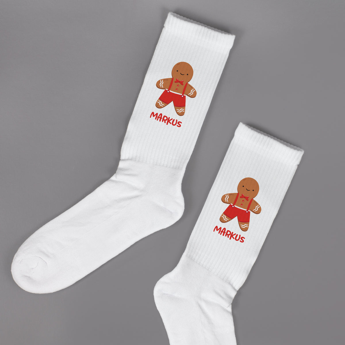 Paar weißer Socken mit Lebkuchenmann-Motiv und personalisiertem Namen, flach auf grauer Oberfläche.