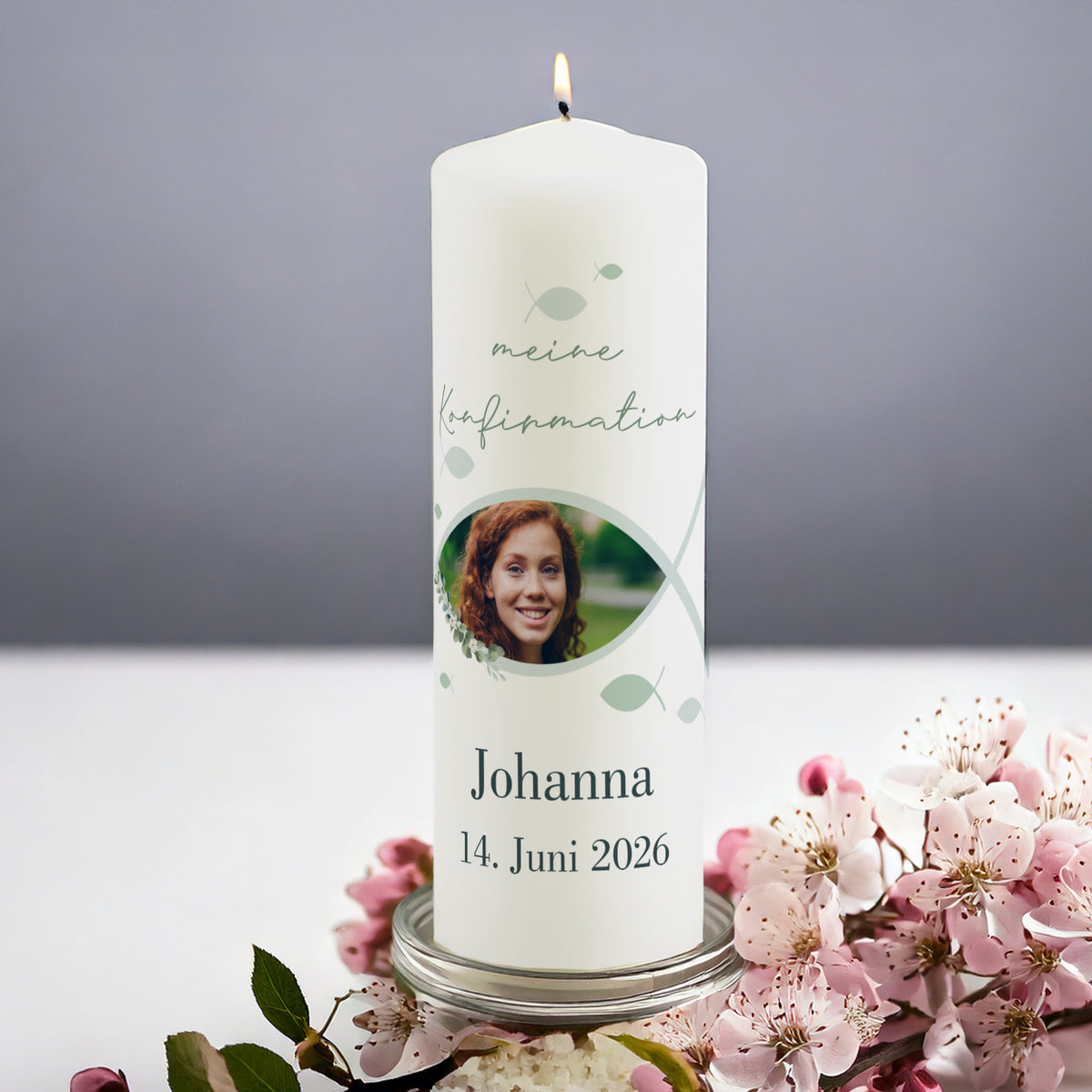 Personalisierte Kerze zur Konfirmation mit Foto. Die weiße Kerze zeigt den Schriftzug „meine Konfirmation“, ein christliches Fischsymbol mit integriertem Foto eines Mädchens und darunter Name „Johanna“ mit Datum „14. Juni 2026“. Die Kerze steht angezündet auf einem Glasteller, umgeben von rosa Blüten.