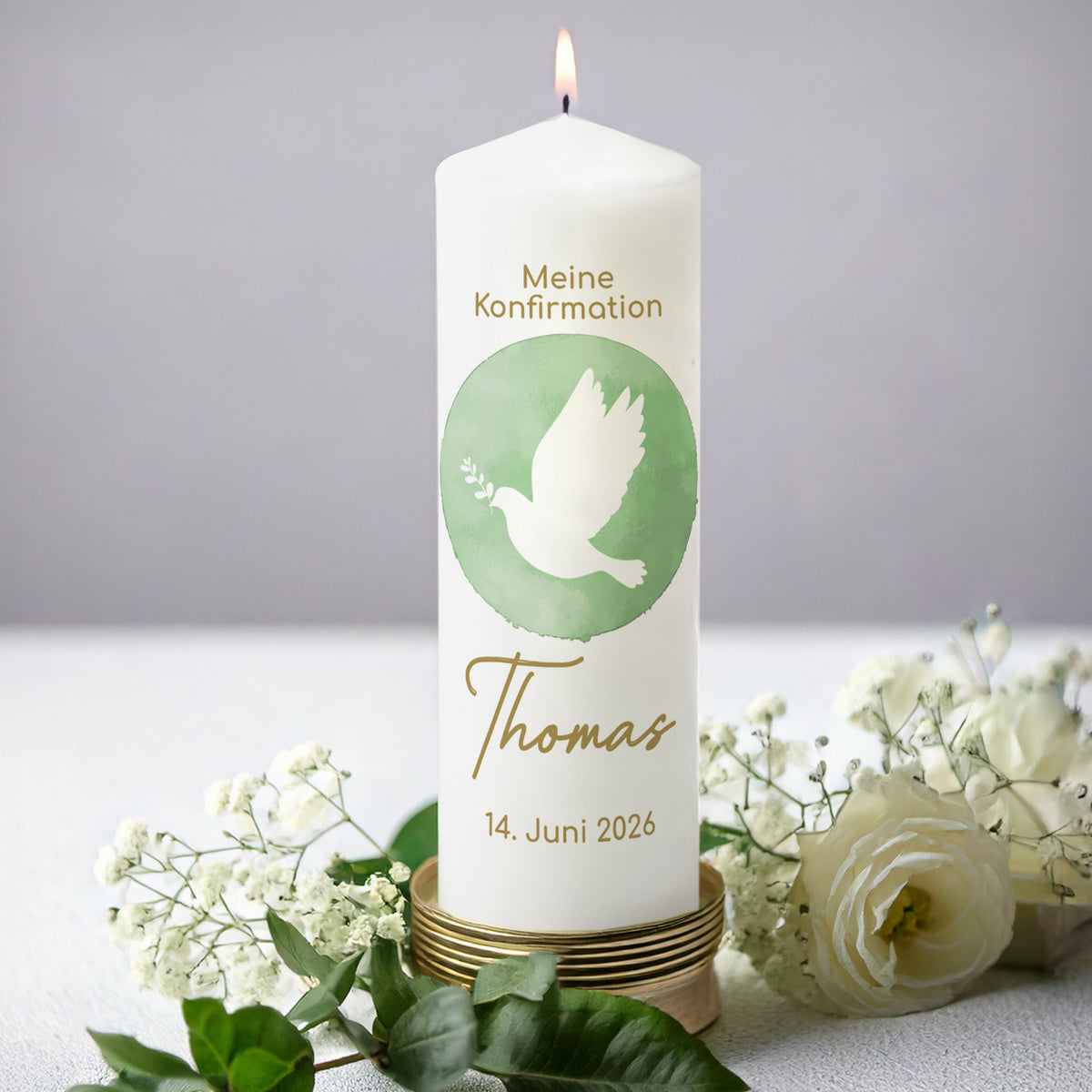Personalisierte Kerze zur Konfirmation mit grünem Aquarellmotiv einer Taube mit Zweig. Der Name "Thomas" und das Datum "14. Juni 2026" sind in goldener Schrift ergänzt. Die festliche Kerze steht angezündet auf einem dekorativen Kerzenhalter, umgeben von weißen Blüten und grünen Blättern, vor einem dezenten grauen Hintergrund.
